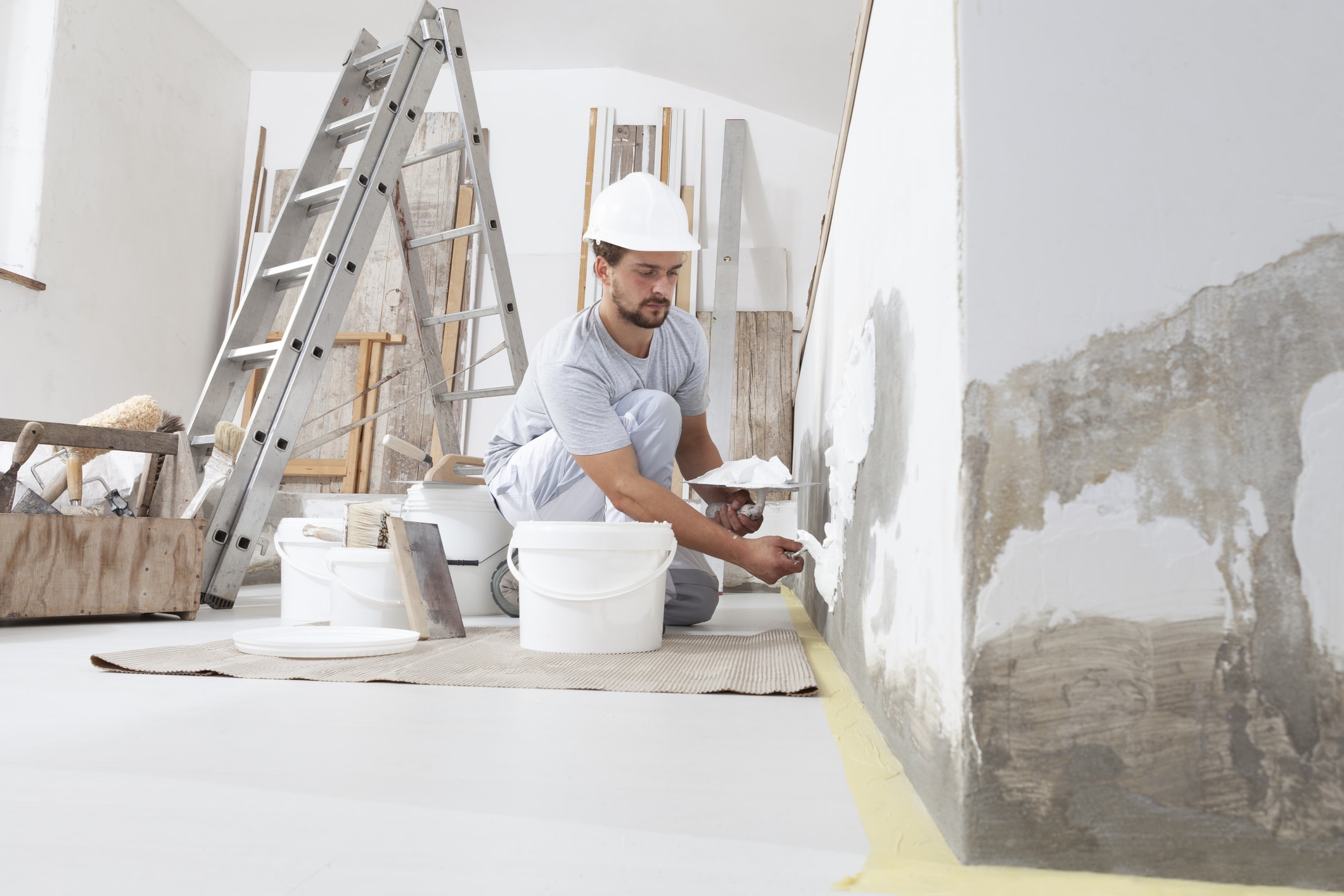 man plasterer construction worker at work, takes plaster from bucket and puts it on trowel to plastering the wall, wears helmet inside the building site of a house